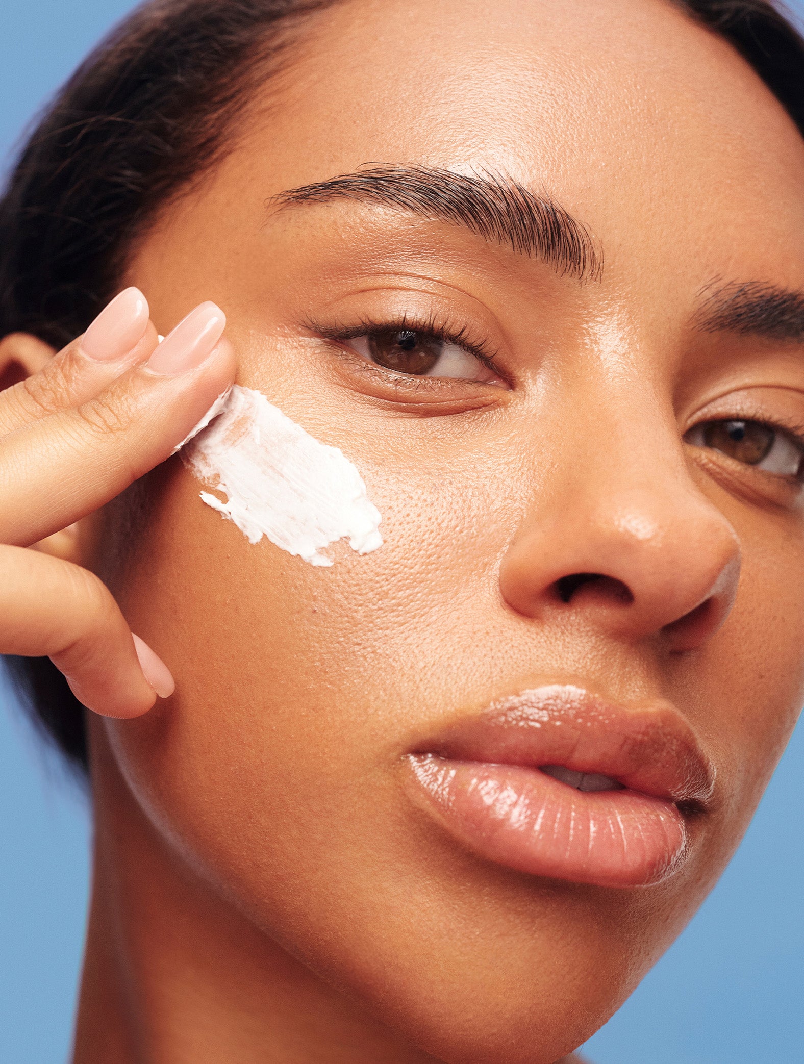 Model applying Lanolips Face Base Day Cream Lanolin Vitamin E to their face, against a blue background. 
